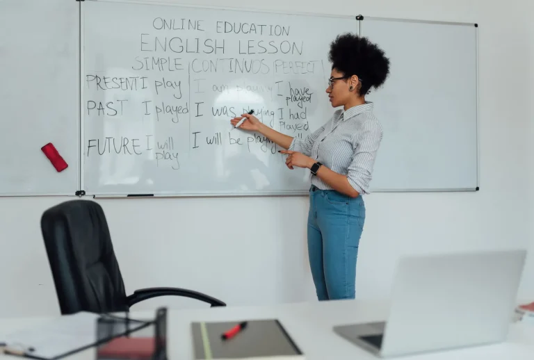 Une enseignante afro-américaine donnant un cours d'anglais langue seconde devant un tableau blanc. Elle explique la grammaire et le vocabulaire anglais, démontrant une pédagogie innovante pour l'enseignement des langues étrangères.